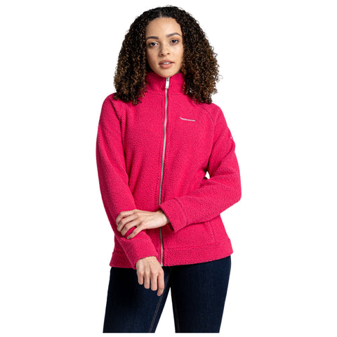 A woman is standing with her arms crossed wearing a bright pink fleece jacket and dark jeans in a neutral background. She has curly hair and a neutral expression.