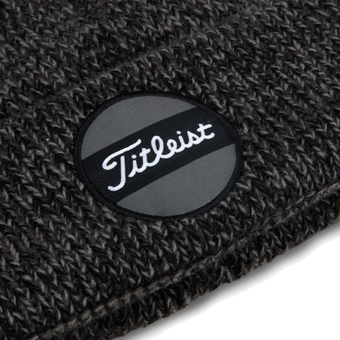 A dark gray knit beanie features a round black patch with the word Titleist in white cursive text and a horizontal gray stripe, creating a casual warm accessory for outdoor wear.