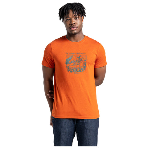 A young man stands wearing an orange t-shirt featuring a graphic design on the back that reads THE JOURNEY IS THE DESTINATION. He has short, curly hair and a neutral expression.