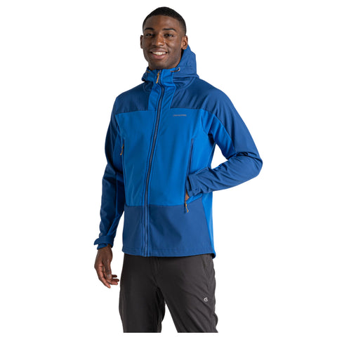 A young man is wearing a blue zip-up jacket with a hood while smiling and standing with one hand in his pocket against a plain white background.