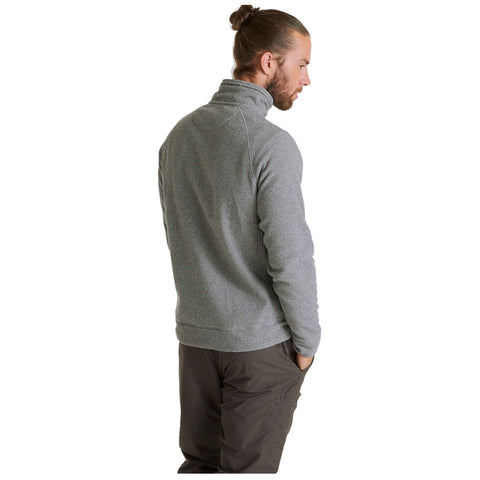 A man with long hair in a bun stands with his back to the viewer wearing a gray pullover and dark pants with his hands in his pockets in a plain background.